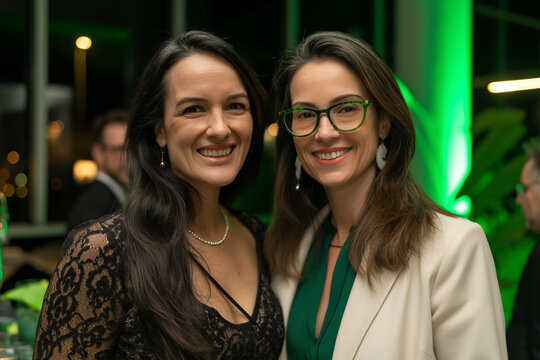 Two women, one with long black hair and the other wearing glasses, posed for photos at an event. The scene was set against green LED lights and sleek architecture.