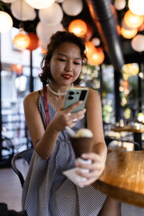 Young woman enjoying ice cream and browsing smartphone in a modern cafe with colorful hanging lights creating a vibrant atmosphere