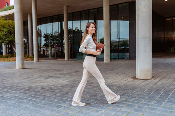 Beautiful smiling businesswoman walking outdoors, holds take away coffee and digital tablet. Female business professional commuter walks on the street in front of office building.