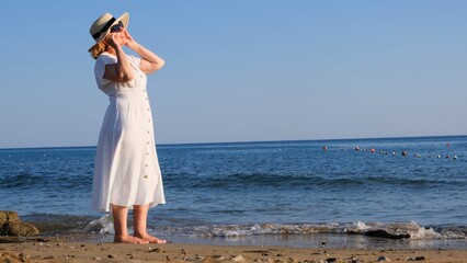 woman in a straw hat and white dress walks along the seashore puts on sunglasses and admires the blue sea on a sunny summer day, enjoying freedom and relaxation. The concept of a senior citizen