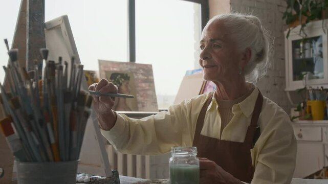 Cheerful woman painting on canvas using brush and gouache colours while attending art class for seniors