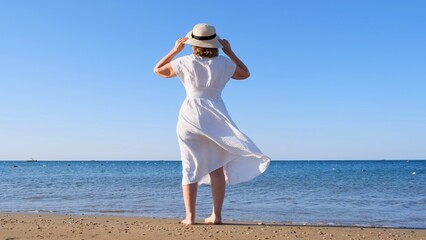 back view on a mature woman in a straw hat and white dress walks along the blue sea coast on a sunny summer day, enjoying freedom and relaxation. The concept of a typical life of pensioners.