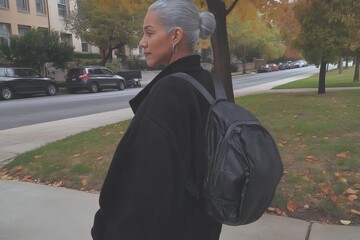 Fototapeta premium A woman wearing a black jacket and a backpack is walking down a street. The scene is set in a residential area with cars parked along the street. The woman is in a hurry