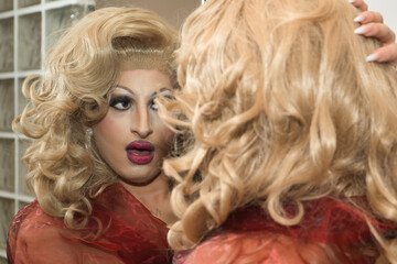 Portrait of drag queen, blonde, attractive, wearing a red chiffon dress, looking flirtatiously in a mirror in her bedroom. Concept pride, diversity, identity, artist.