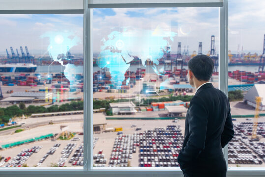 A businessman stands by a window overlooking a port, viewing digital world map projections and global trade logistics data.