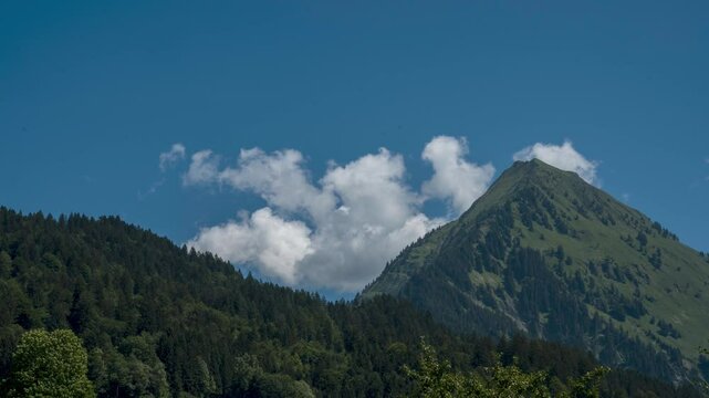 Das Zeitraffer-Video zeigt eine malerische Berglandschaft an einem klaren, sonnigen Tag. Ein gr&uuml;ner, bewaldeter H&uuml;gel im Vordergrund und ein steiler Berggipfel, der sich in den blauen Himmel erhebt.