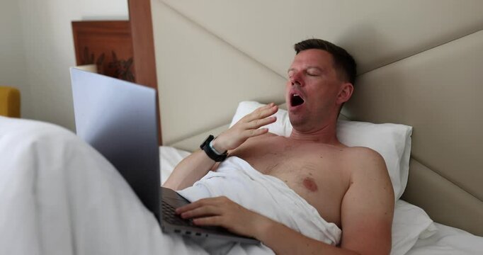 Young man suffers from lack of sleep with laptop in bedroom. Freelancer overwork and overtime