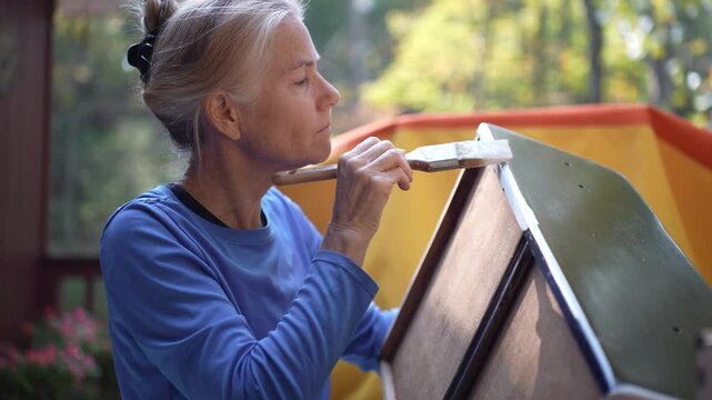 A woman with gray hair, wearing a blue shirt, paints a wooden object with white paint using a brush. The background is blurred with a yellow and orange object and green foliage.