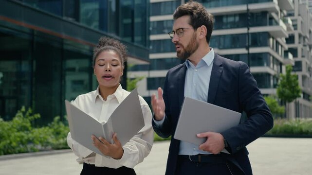 Two angry negative arguing talking multiracial business colleagues walk in city African woman businesswoman female girl quarrel discuss data work problem Caucasian man businessman guy male hold laptop