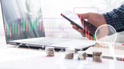 A person is holding a cell phone in front of a laptop with a graph on the screen. The graph shows a series of numbers and the person appears to be looking at it