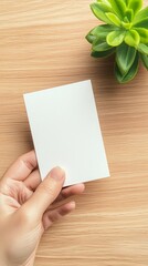 Obraz premium A hand holds a blank card beside a small green plant on a wooden surface, creating a calm and minimalist aesthetic.
