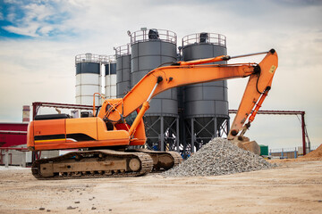 Excavator moving gravel at a busy construction site with silos in the background during a clear day © Anoo