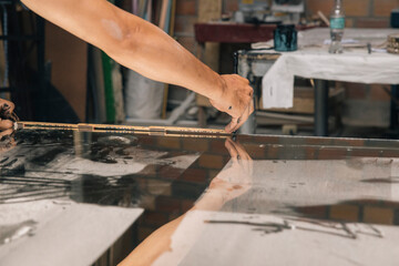 An artisan hands use a glass cutter to trace a line across a sheet of glass, preparing it for breaking in a workshop setting