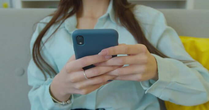 Beautiful young woman holding a smartphone in hands and typing on its screen while chatting in the living room at home. Indoors. Close-up.