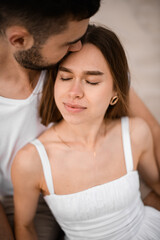 Young woman sits on the sand in the arms of her fiance with her eyes closed and her head resting on his shoulder