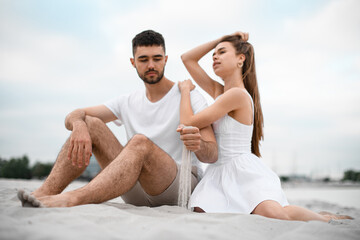 Fototapeta premium Young couple sitting on the sand on the seashore. A guy pours sand through his fingers while his girlfriend leans on his shoulder