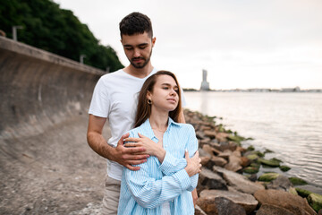 Young couple in love are standing on a stony river bank, the man hugs the woman from behind