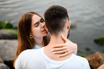 Fototapeta premium Young pretty woman kisses her fiance's ear and hugs his neck, who is sitting with his back