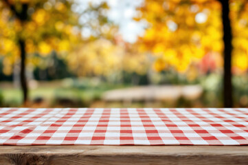 Red and white checkered tablecloth on a wooden picnic table with blurred autumn foliage in the background. Outdoor dining and fall season concept