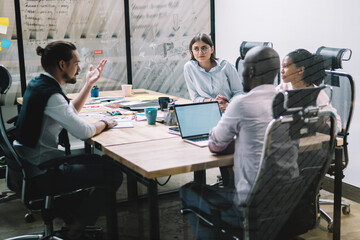 Intelligent multicultural people cooperating on ideas for financial strategy of company during brainstorming meeting in conference room, serious partners sitting at desktop and consultancy together