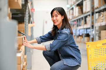 Woman squatting and selecting a box from a shelf in a warehouse, smiling and focused.