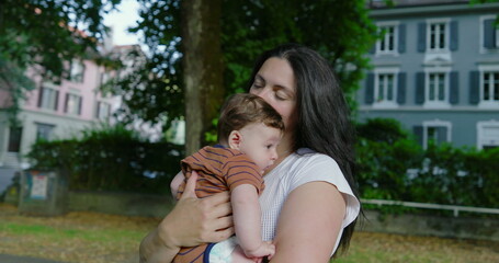 Mother holding her baby on a tree-lined urban path, gazing lovingly at her child, surrounded by buildings and greenery, with a calm and nurturing atmosphere captured