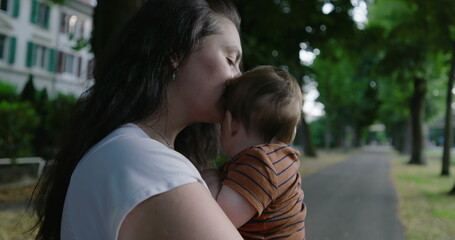 Fototapeta premium Mother kissing her baby’s head while holding him close, standing on a tree-lined path, expressing tenderness and love, with a peaceful urban setting