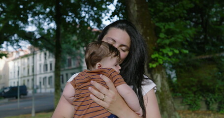 Fototapeta premium Mother smiling warmly while holding her baby on a tree-lined urban path, showcasing a moment of affection and tenderness, surrounded by greenery and residential buildings
