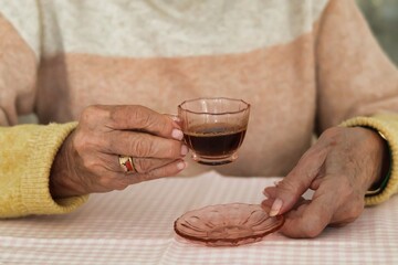 Elderly woman's hands holding a small pink cup of coffee; pastel colors - yellow and pink