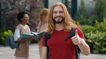 Happy Caucasian student guy man male schoolboy in city outdoors university college campus showing thumbs up to camera smile enjoy education study knowledge generation Z students talking on background