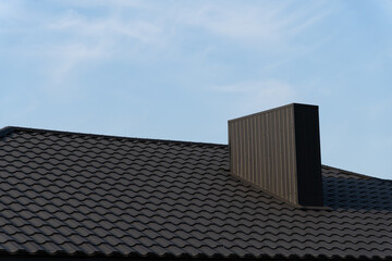 The image features a sleek, modern tiled roof of a newly built house, showcasing a prominent chimney against a bright blue sky. The clean lines emphasize contemporary architecture