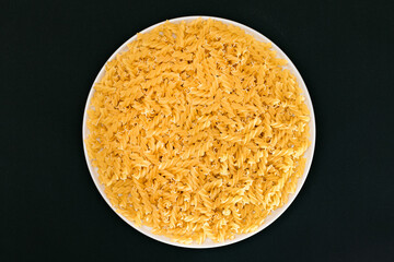 Uncooked Fusilli Pasta Lying on White Plate on Black Background. Raw and Dry Macaroni. Unhealthy and Fat Food - Top View