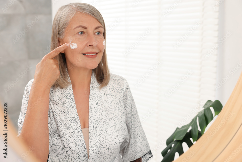 Poster Senior woman applying face cream near mirror at home