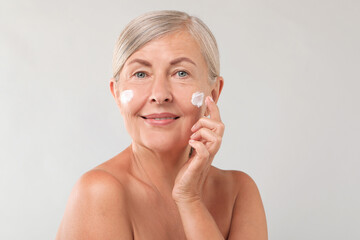 Senior woman applying face cream on light background