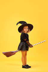 Cute girl with broom dressed like witch on yellow background. Halloween celebration