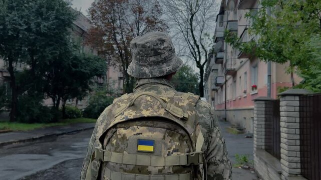 A Ukrainian soldier in a military panama hat and with a khaki backpack walks down the street. The soldier returns from war in the rain.