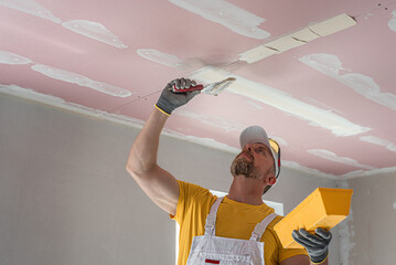 The worker make a plasterboard ceiling. He does taping plaster drywall ceiling joints. He is...