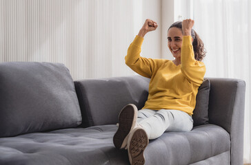 Obraz premium Portrait latin american woman one person smile enjoy sitting sofa and looking television raise your hand, happy in choosing to watch series on most wonderful vacation day in house living room