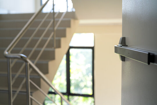Fire exit metal door in opening position with background of the stairway with railing handle, using for emergency escape route. Building interior part photo, selective focus.