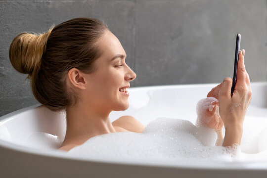 Happy young cheerful brunette woman relaxing in foamy bath with smartphone cell phone reading screen - Powered by Adobe