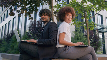 Two people multiracial colleagues diversity business partners working outdoors on bench with two laptops African woman Indian Arabian man work together with computers back to back smiling at camera