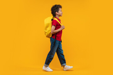 Full length studio portrait of a cute casually dressed happy smiling confident African American schoolboy walking with his bright colored backpack over orange yellow background