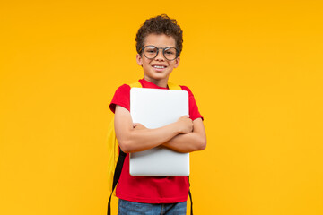 Portrait of cute happy smiling casually dressed African American schoolboy posing in studio over yellow background with a laptop in his hands