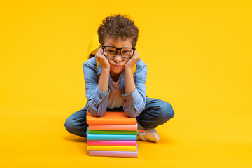 Cute little African American kid student leaning on the books pile face buried in his hands looking...