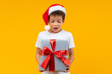 Cute little African American boy in basic white t-shirt and Santa Claus hat looking at the Christmas present box in his hands with surprised face expression, isolated on yellow background