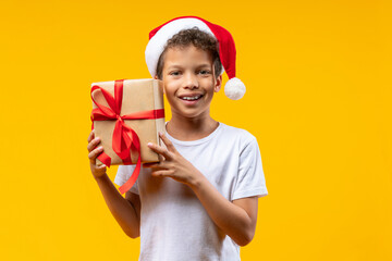 Portait of joyful African American kid wearing Santa Claus hat posing in studio over bright colored yellow background holding a gift box wrapped in kraft paper and decorated with red satin ribbon