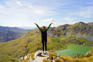 A girl photographed from behind stands with her arms outstretched joyfully on top of a cliff against a beautiful mountain landscape. A female tourist trekking near Kapetanovo Lake, Montenegro.