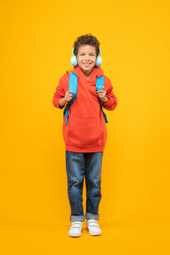 Vertical shot of cute casually dressed Afrcan American schoolboy smiling to the camera and posing on yellow background with his backpack and wireless headphones