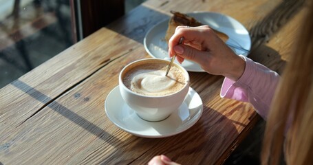 Woman guest stirs creamy coffee with spoon in coffeeshop lit by sunlight closeup. Lady orders caffeine drink with cake at rustic wooden counter. Morning vibes
