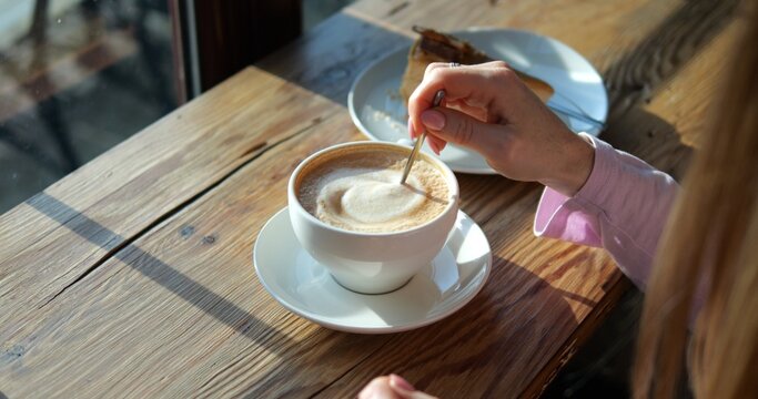 Woman guest stirs creamy coffee with spoon in coffeeshop lit by sunlight closeup. Lady orders caffeine drink with cake at rustic wooden counter. Morning vibes